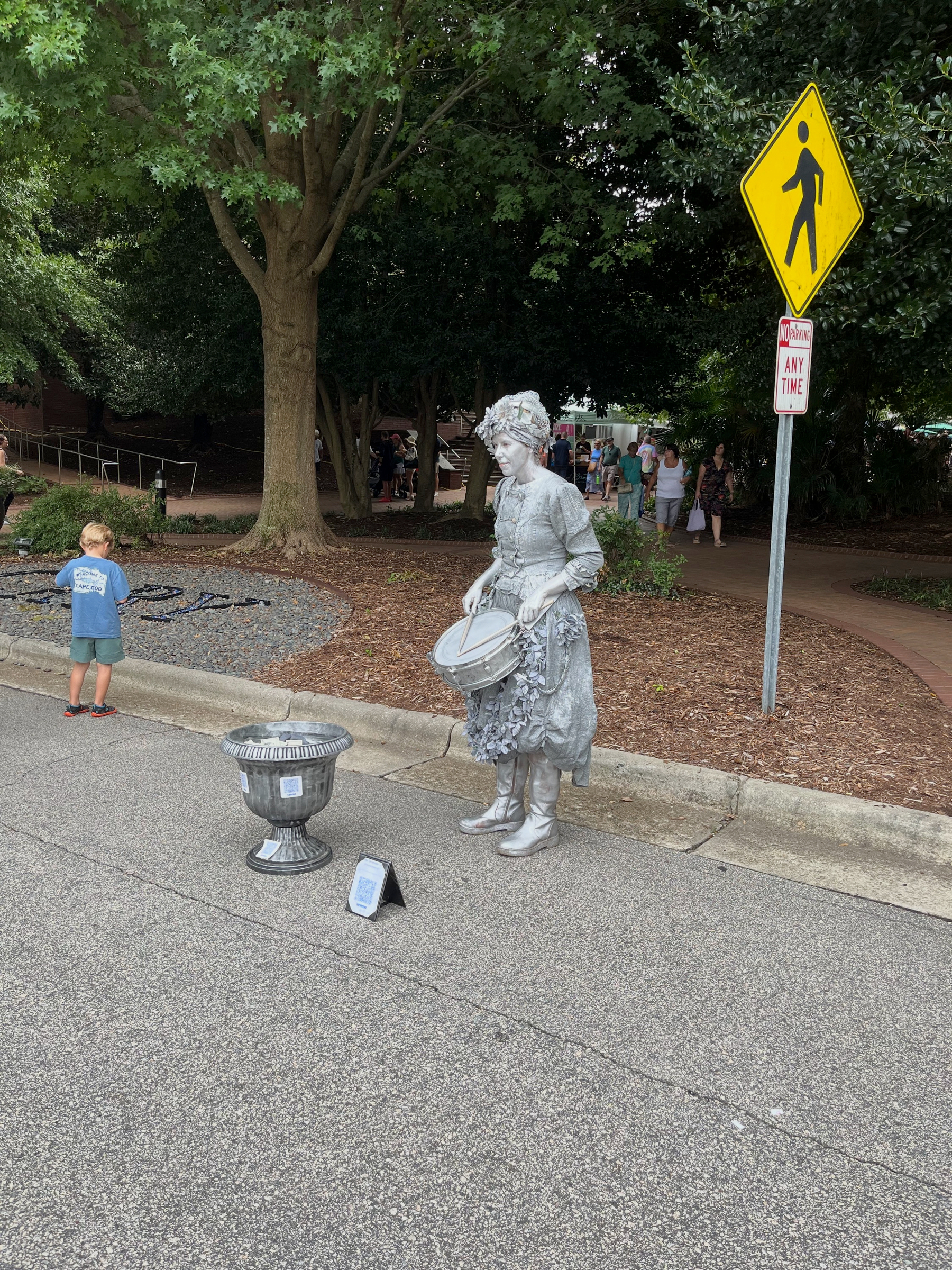 A street performer dressed and painted like a silver statue is standing on the sidewalk playing a drum, with a small container placed in front for tips.
